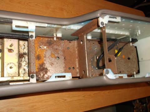 1999 Mercury Grand Marquis - The image shows the interior of a vehicle's center console area, revealing rusted metal brackets and wiring. The console's gray trim is vis…