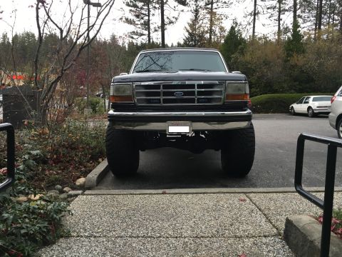 1993 Ford Bronco - The front view of a lifted black Ford Bronco with large tires is parked on a paved surface. The vehicle is facing the camera, with trees an…