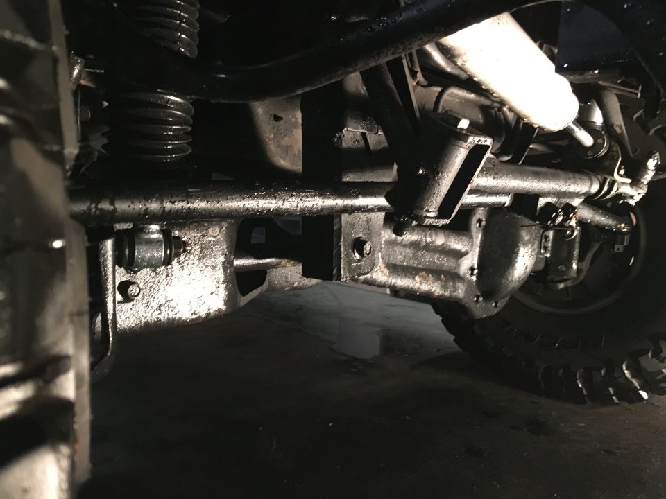 Underside of a vehicle showing front axle, steering, and suspension components coated in grease.