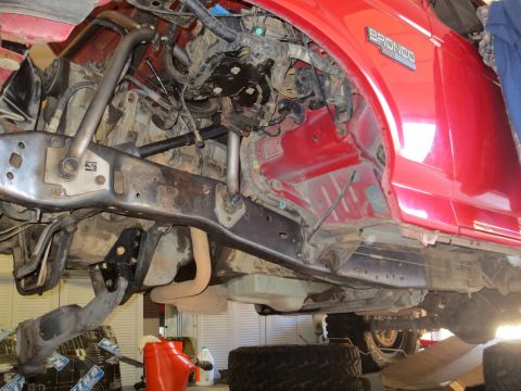 1996 Ford Bronco - Underside view of a red 1996 Ford Bronco with suspension modifications, showing the frame, exhaust, and part of the drivetrain.