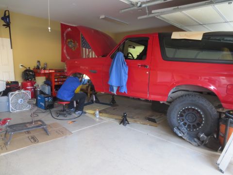 1996 Ford Bronco - A person is working on the front wheel well of a red Ford Bronco in a garage. The vehicle is raised on jack stands, and tools and equipment…
