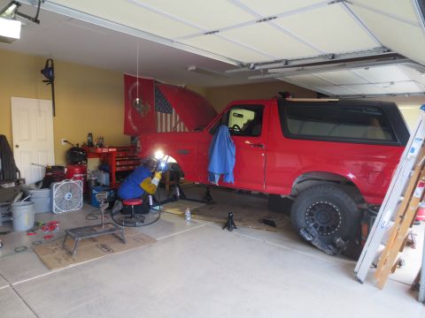 1996 Ford Bronco - A person in welding gear is working on the undercarriage of a red Ford Bronco in a garage. The vehicle is raised on jack stands, and tools…