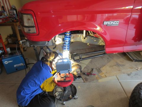 1996 Ford Bronco - A person wearing a welding mask and protective gear is welding the suspension of a red Ford Bronco. A Miller welding machine is visible to…