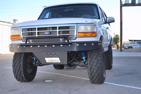1996 Ford Bronco - Front view of a lifted, silver 1996 Ford Bronco with an aftermarket black bumper and large off-road tires. Blue coilover shocks are visible…