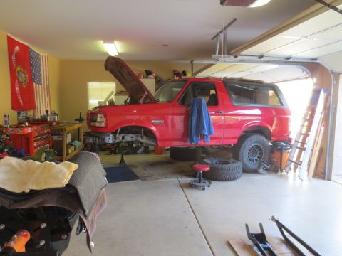 1996 Ford Bronco - A red 1996 Ford Bronco is parked in a garage with its hood open and front end disassembled. The vehicle is on jack stands, and various tool…