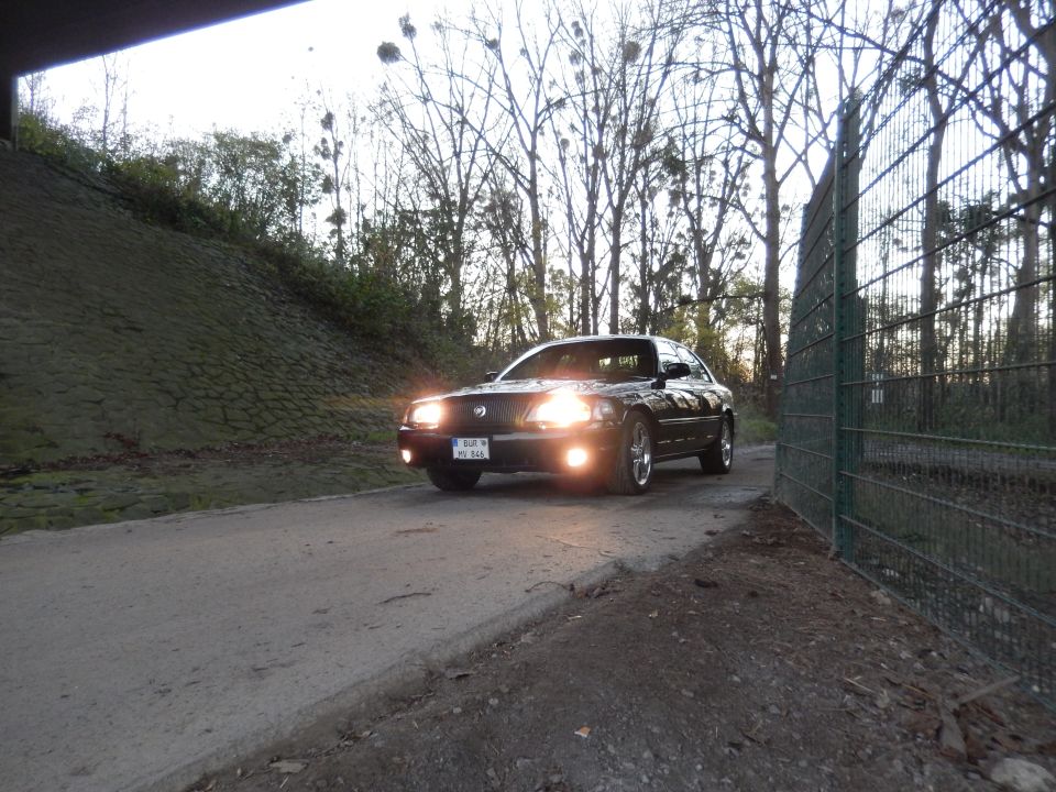 Black 2003 Mercury Marauder with headlights on, parked on a road next to a fence.