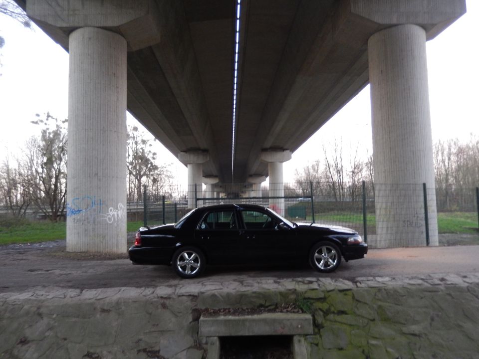 Black sedan parked under a concrete overpass, side view.