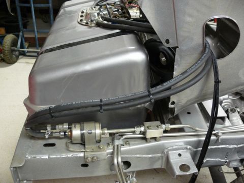 Gray fuel tank with black fuel lines and a fuel filter mounted to a vehicle chassis.