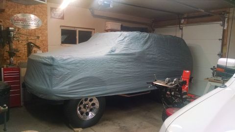 1994 Ford Bronco - A large vehicle, likely an SUV, is covered by a grey tarp in a garage. The vehicle has large chrome wheels and appears to be a 1994 Ford Br…