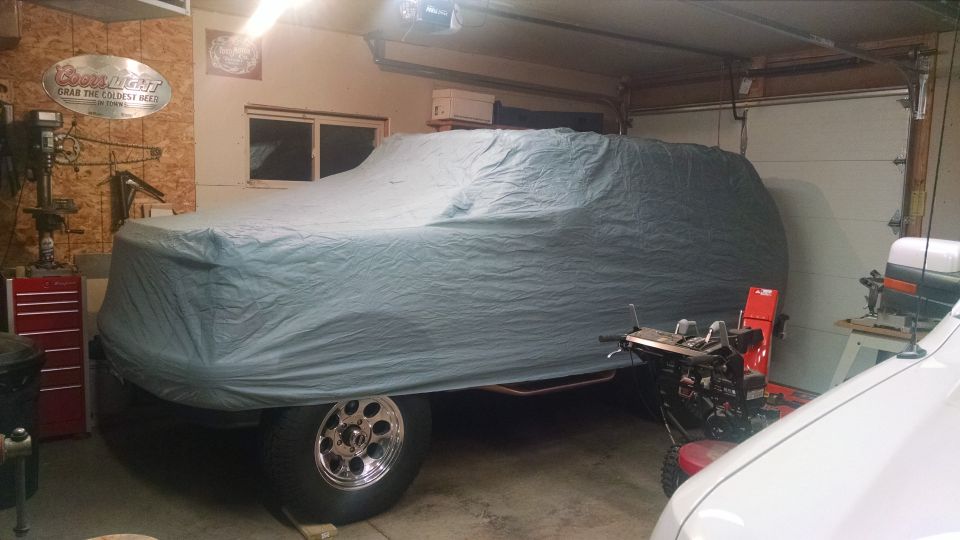 1994 Ford Bronco covered by a grey tarp in a garage, showing chrome wheels.