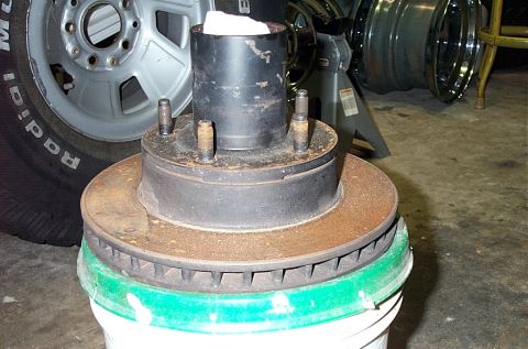 1995 Ford Bronco - A vented front brake rotor still attached to its wheel hub, sitting upright on a white five-gallon bucket in a garage, with the wheel studs…