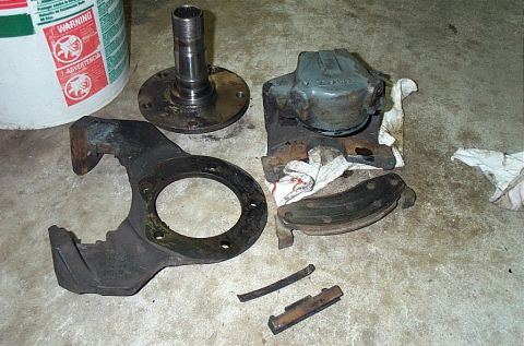 1995 Ford Bronco - Disassembled front axle and brake hardware laid out on a concrete garage floor: a splined spindle on its flange, a cast brake caliper ancho…