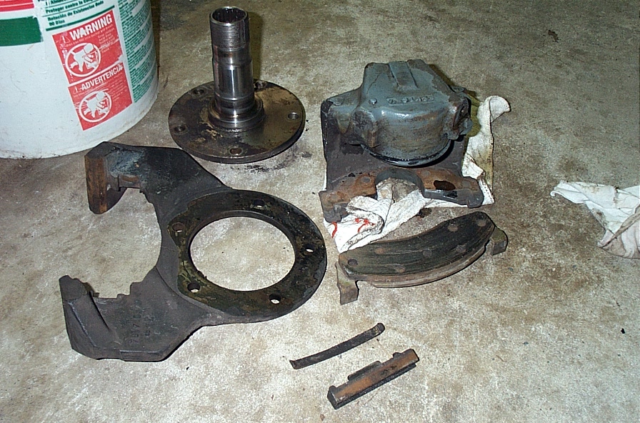Disassembled axle spindle, caliper and brake dust shield laid out on a garage floor