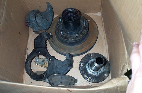 1995 Ford Bronco - A cardboard box holds a set of used front brake and hub components: a rusty rotor with an integral five-lug hub, a cast steering knuckle, a…