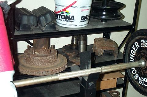 1995 Ford Bronco - Rusty front axle parts stored on a black steel garage shelf — a brake rotor still mounted on its hub and spindle, a second spindle on a bar…