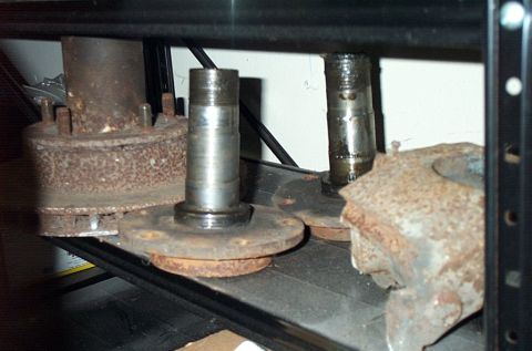 1995 Ford Bronco - Close-up of two used front axle spindles bolted to their flanges sitting on a black metal shelf, their machined bearing journals showing we…