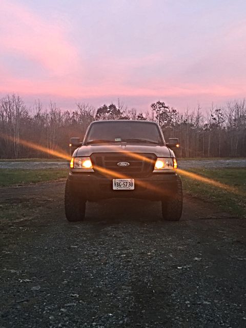 1991 Ford Bronco - A dark-colored Ford pickup truck is parked on a gravel lot with its headlights on, facing the camera. The sky behind it is a gradient of pi…