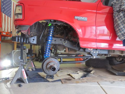 1996 Ford Bronco - The undercarriage of a red Ford Bronco is shown with the front suspension disassembled, featuring a bright blue coilover shock and a large…