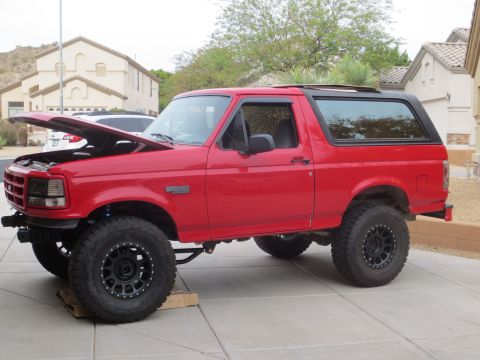 1996 Ford Bronco - A red 1996 Ford Bronco is parked on a paved driveway with its hood open. The SUV has large, black aftermarket wheels and tires.