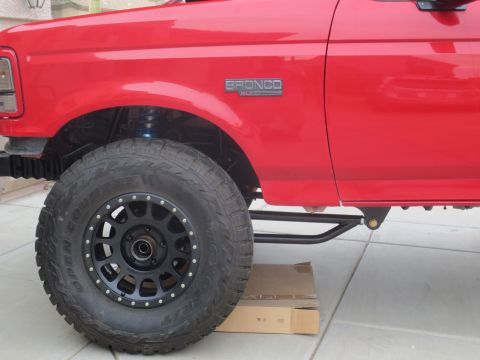 1996 Ford Bronco - Close-up side view of a red Ford Bronco's front fender and wheel. The vehicle features a black aftermarket wheel and tire, and a black side…