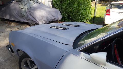 1976 Ford Ranchero - A light blue primer-coated Ford Ranchero hood is visible, featuring two rectangular chrome-trimmed vents. Another vehicle covered by a grey…