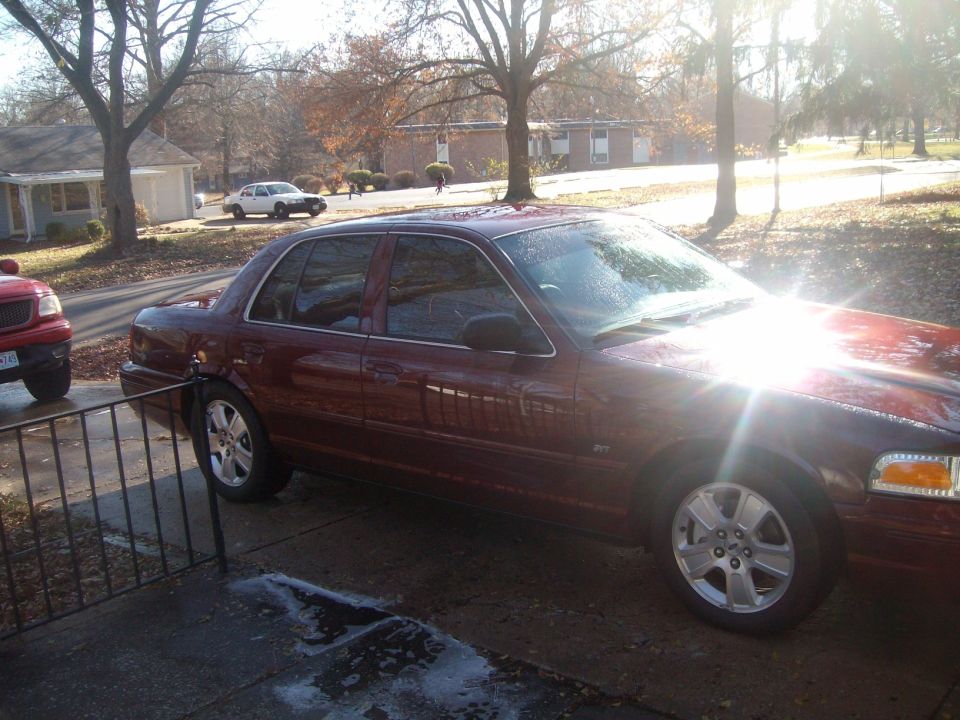 Dark red sedan parked on a driveway with a red truck to the left, sun glare on windshield.