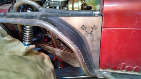 1985 Jeep CJ7 - A custom metal fender flare with a skull and piston emblem is attached to the side of a red Jeep CJ7. The vehicle features exposed suspensi…