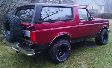 1994 Ford Bronco - The rear passenger side of a dark red 1994 Ford Bronco is shown parked on a grassy lawn. A spare tire is mounted on the rear swing-away car…