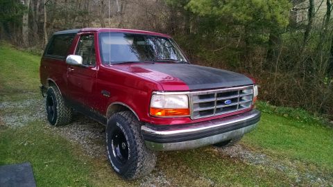 1994 Ford Bronco - A red 1994 Ford Bronco with a black hood sits parked on a gravel driveway next to a grassy area with trees in the background.