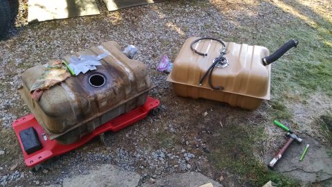 1994 Ford Bronco - Two automotive fuel tanks are shown outdoors on gravel and grass. The tank on the left is rusty and sits on a red creeper, while the tank o…