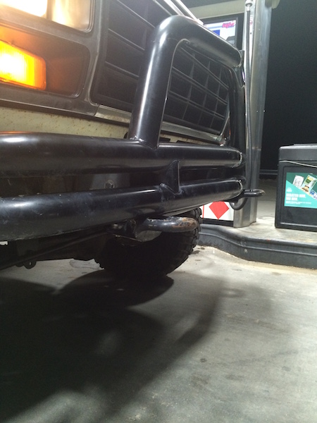 1981 Ford Bronco - A close-up, low-angle shot of the front bumper and grille of a dark-colored vehicle, possibly an older Ford Bronco, at a gas station. The v…