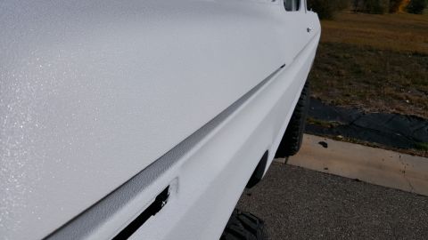1978 Ford Bronco - The side of a white vehicle with a textured finish is shown from a low angle, highlighting the door seam and the rear fender. A portion of…