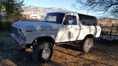 1978 Ford Bronco - A white Ford Bronco is parked outdoors on a dirt road covered with fallen leaves. The vehicle has large off-road tires and a trailer is vis…