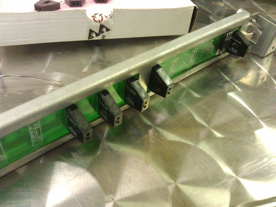 Electronic board with multiple black connectors mounted in a metal housing. Box with black triangular objects in background