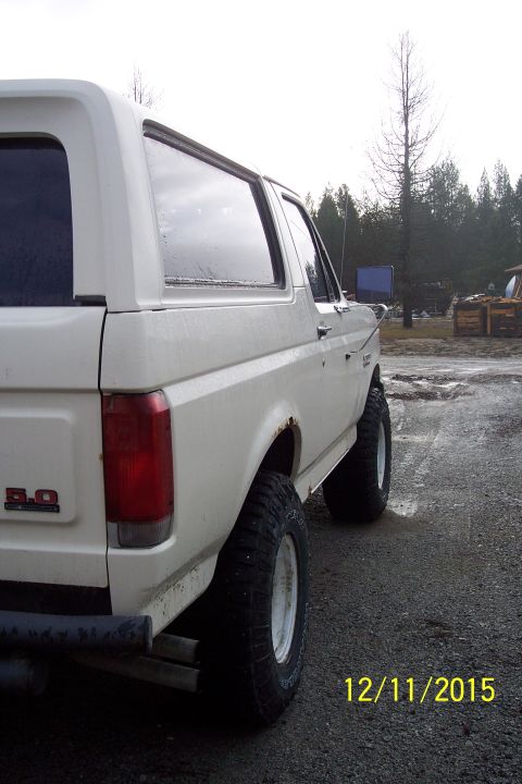 1990 Ford Bronco - A white 1990 Ford Bronco is viewed from the rear quarter panel, showing its lifted suspension and off-road tires.