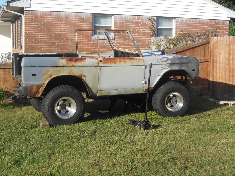 Old Ford Bronco body, rusty and missing parts, sitting in a grassy yard. Flames painted on the fender