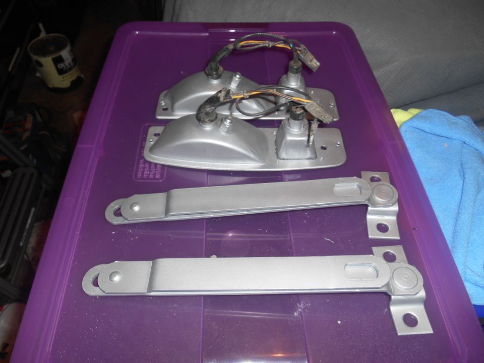 Two silver tail light assemblies with wiring and two silver metal brackets with pivot points, all on a purple plastic container