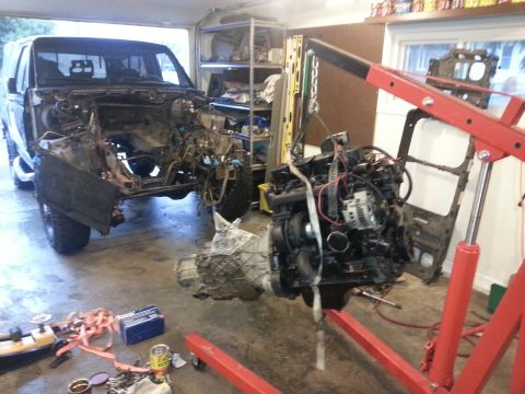 1994 Ford Bronco - A black Ford Bronco is partially disassembled in a garage, with its engine hoisted by a red engine hoist.