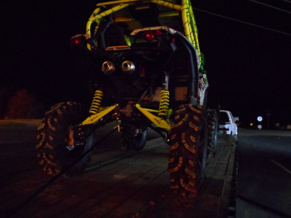 rear view of a yellow off-road buggy with large tires on a trailer, a white pickup truck is visible behind it at night