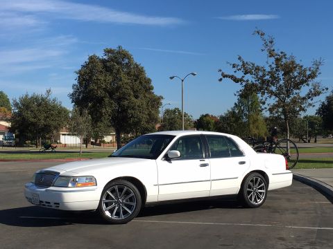 1989 Mazda MX-6 - White sedan parked in a lot under a blue sky with scattered clouds. A bicycle is mounted on the rear.