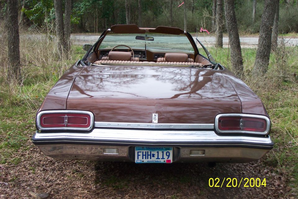 Brown 1970s Oldsmobile convertible seen from the rear, top down, on a wooded track