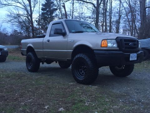 1991 Ford Bronco - A lifted, two-wheel-drive Ford Ranger pickup truck is parked on a gravel and grass area with bare trees in the background. The truck is tan…