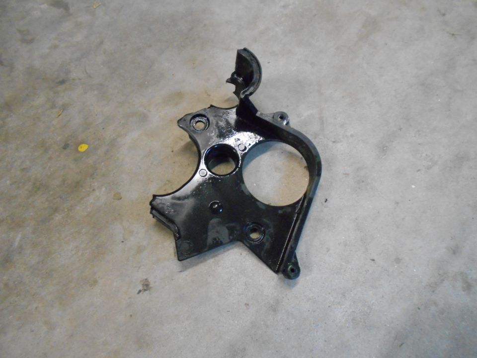 Black metal engine timing cover with circular cutouts on a concrete floor.
