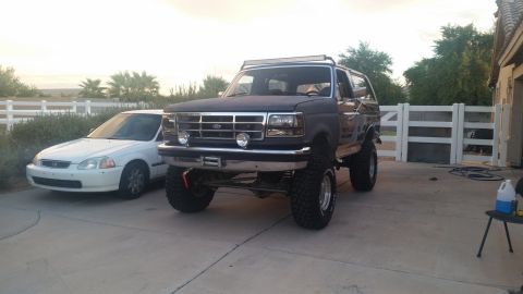 1991 Ford Bronco - A lifted, dark-colored Ford Bronco with a light bar on the roof is parked on a concrete driveway next to a white Honda Civic. The Bronco ha…