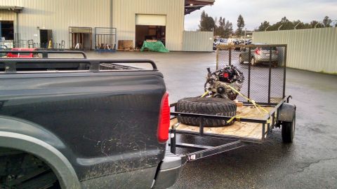 1985 Jeep CJ7 - A dark-colored pickup truck is parked next to a trailer carrying a car engine and a spare tire. The trailer is attached to the truck's hitc…
