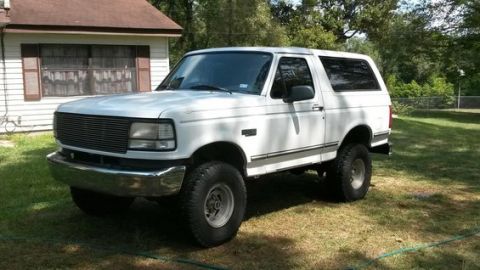 1996 Ford Bronco - A white 1990s Ford Bronco is parked on a grassy lawn in front of a house. The SUV has aftermarket wheels and tires, and a custom grille.