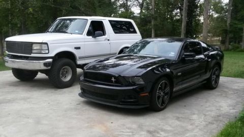 1996 Ford Bronco - A lifted white 1990s Ford Bronco sits next to a black Ford Mustang GT on a concrete driveway. The Bronco has large off-road tires and a chr…