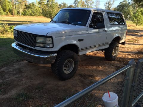 1996 Ford Bronco - A white, two-door Ford Bronco is parked on a dirt lot with a grassy field and trees in the background. The front of the vehicle is facing t…