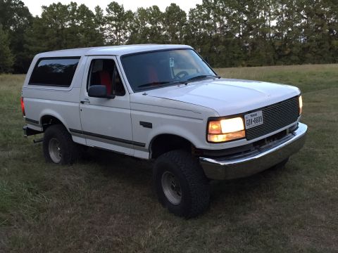 1996 Ford Bronco - A white 1996 Ford Bronco with a chrome grille and aftermarket wheels is parked on a grassy field with trees in the background.