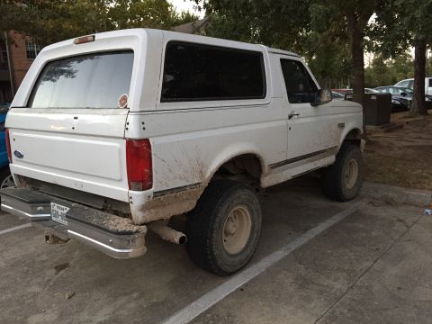 1996 Ford Bronco - The rear driver's side of a muddy white 1996 Ford Bronco is shown parked on pavement. The vehicle has a Texas license plate reading "GOV-86…